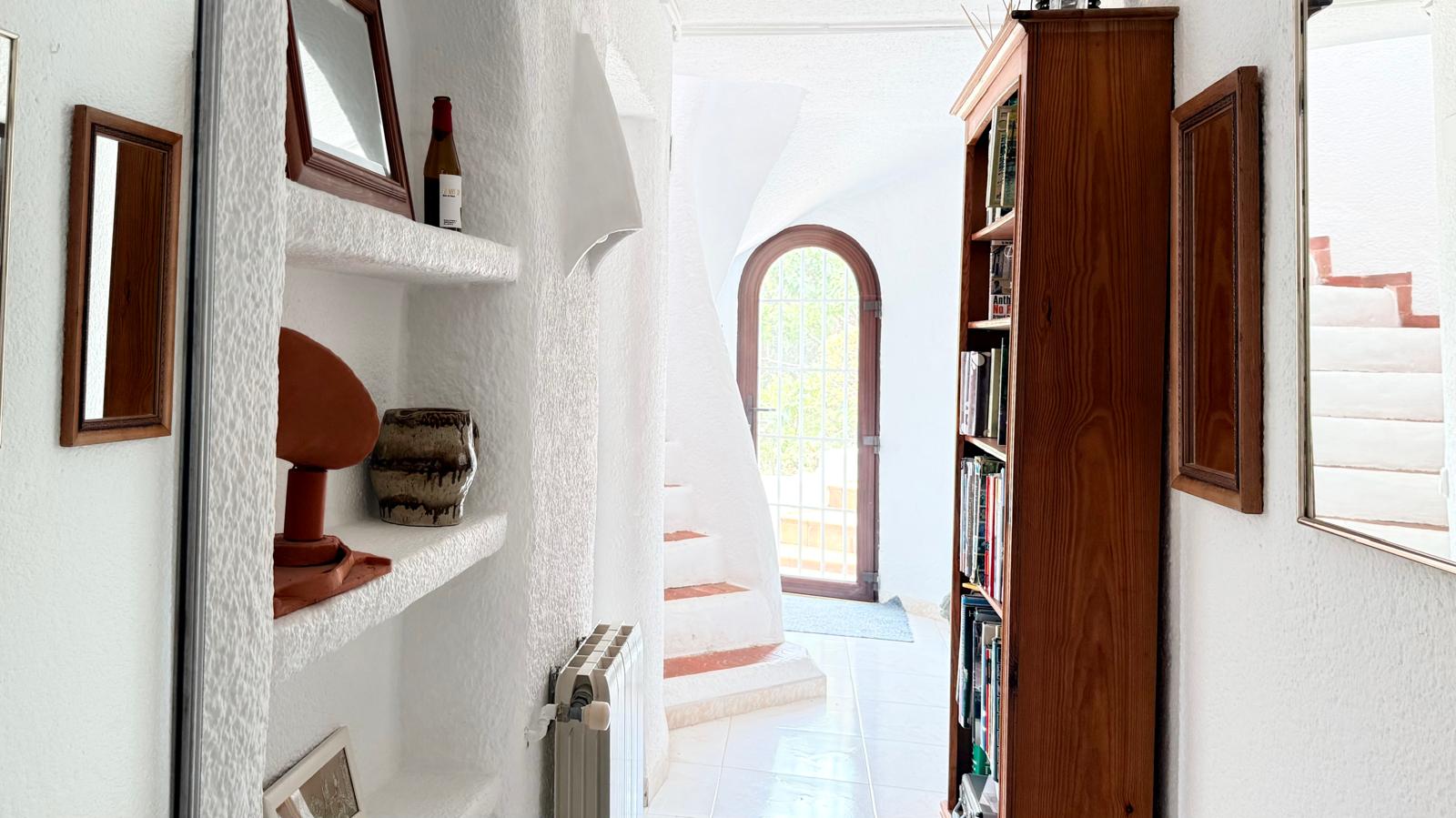 Hal in Ibiza-stijl met witte muren met textuur, ingebouwde nissen en een karakteristieke gebogen trap met terracotta details die naar de bovenverdieping leidt.