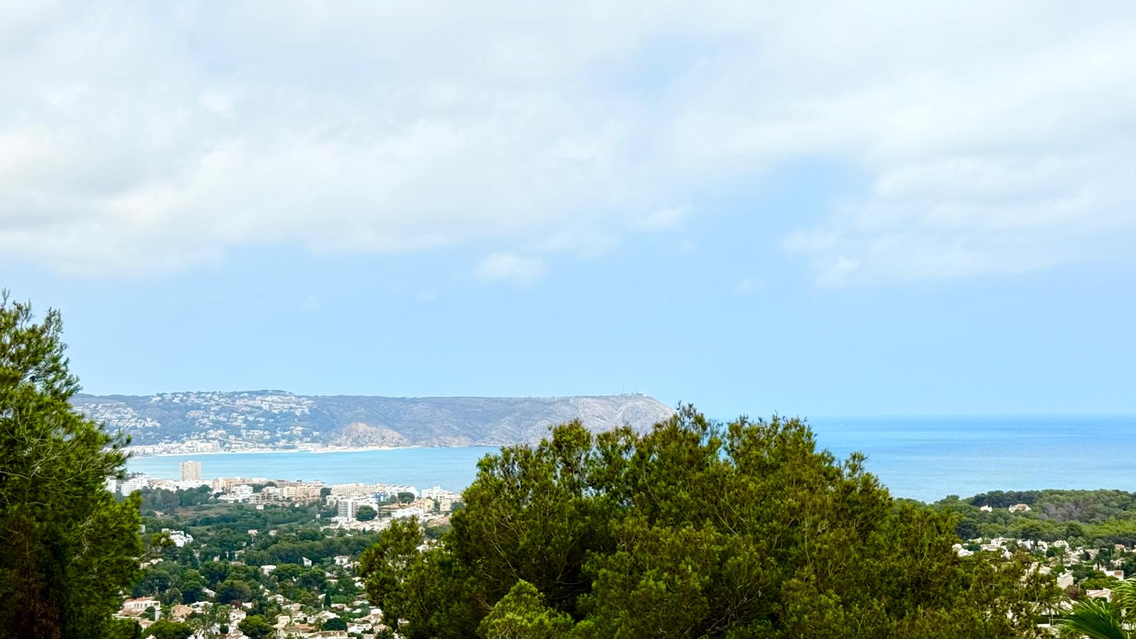 Panoramisch uitzicht op de baai van Jávea en de Middellandse Zee, met de Cap de Sant Antoni aan de horizon, omringd door volwassen pijnbomen op het perceel.