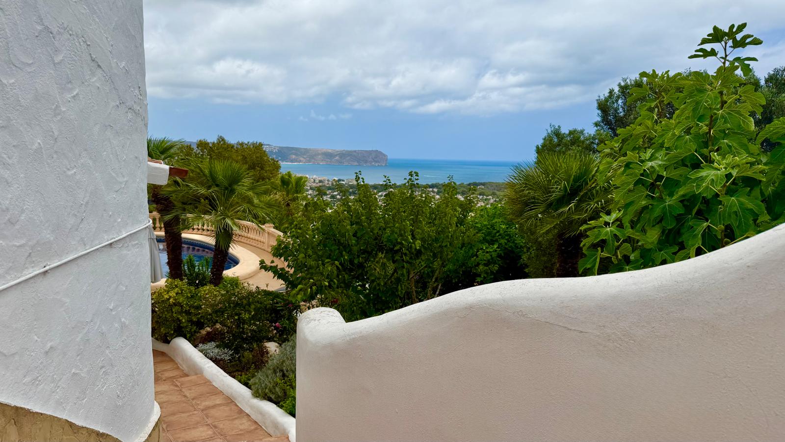 Panoramisch uitzicht op zee over de baai van Jávea vanaf de buitentrap, met witgekalkte gebogen muren en een volgroeide mediterrane tuin met palmbomen.