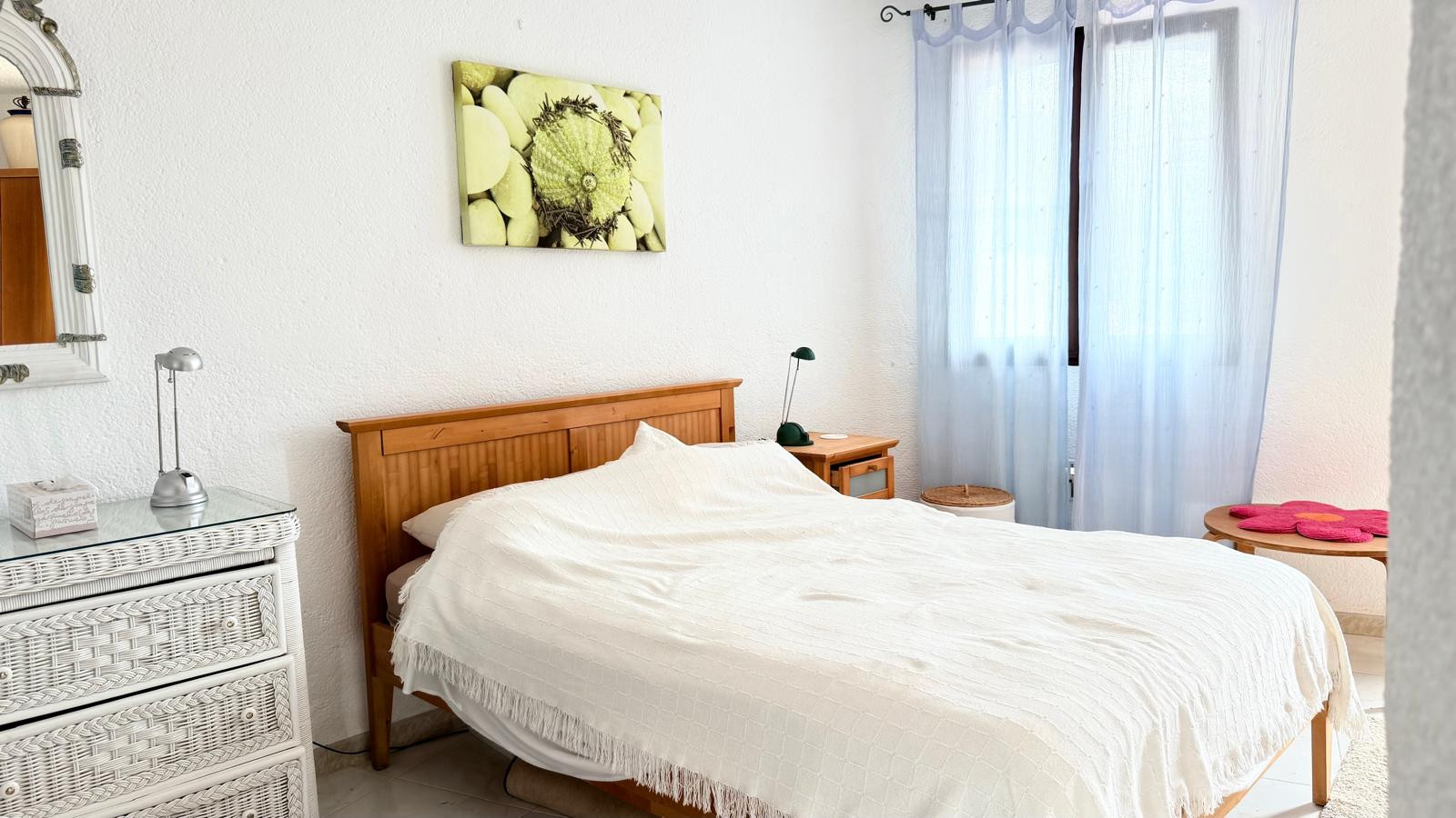 Slaapkamer in mediterrane stijl met witte muren met textuur, een houten bed, witte rieten ladekast en lichtblauwe gordijnen die het daglicht filteren.