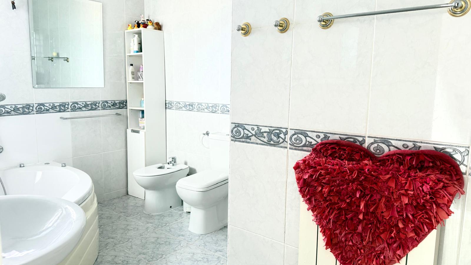 Volledige badkamer met hoek-hydromassagebad, wit sanitair en betegelde wanden met een decoratieve rand. Voorzien van een bidet en een hoge witte opbergkast.
