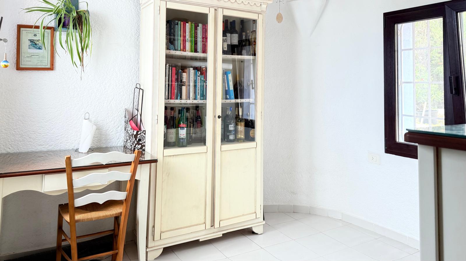 Werkhoek met een wit houten bureau, rustieke stoel en een klassieke vitrinekast voor boeken en flessen op een lichte tegelvloer.