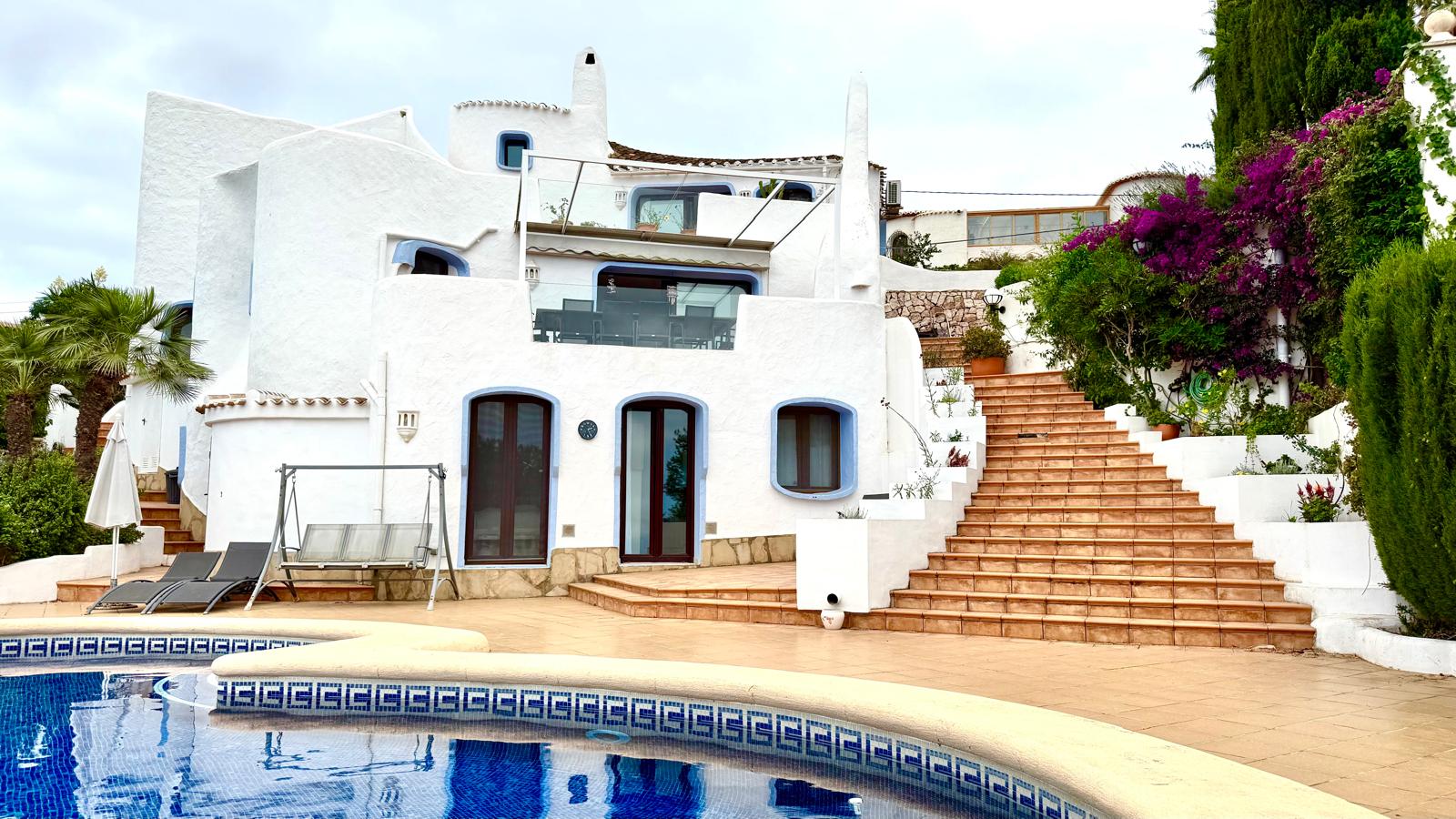 Witte gevel in Ibiza-stijl met blauwe details, een ruim terracotta terras met brede trap en een privézwembad omgeven door volwassen mediterrane vegetatie.