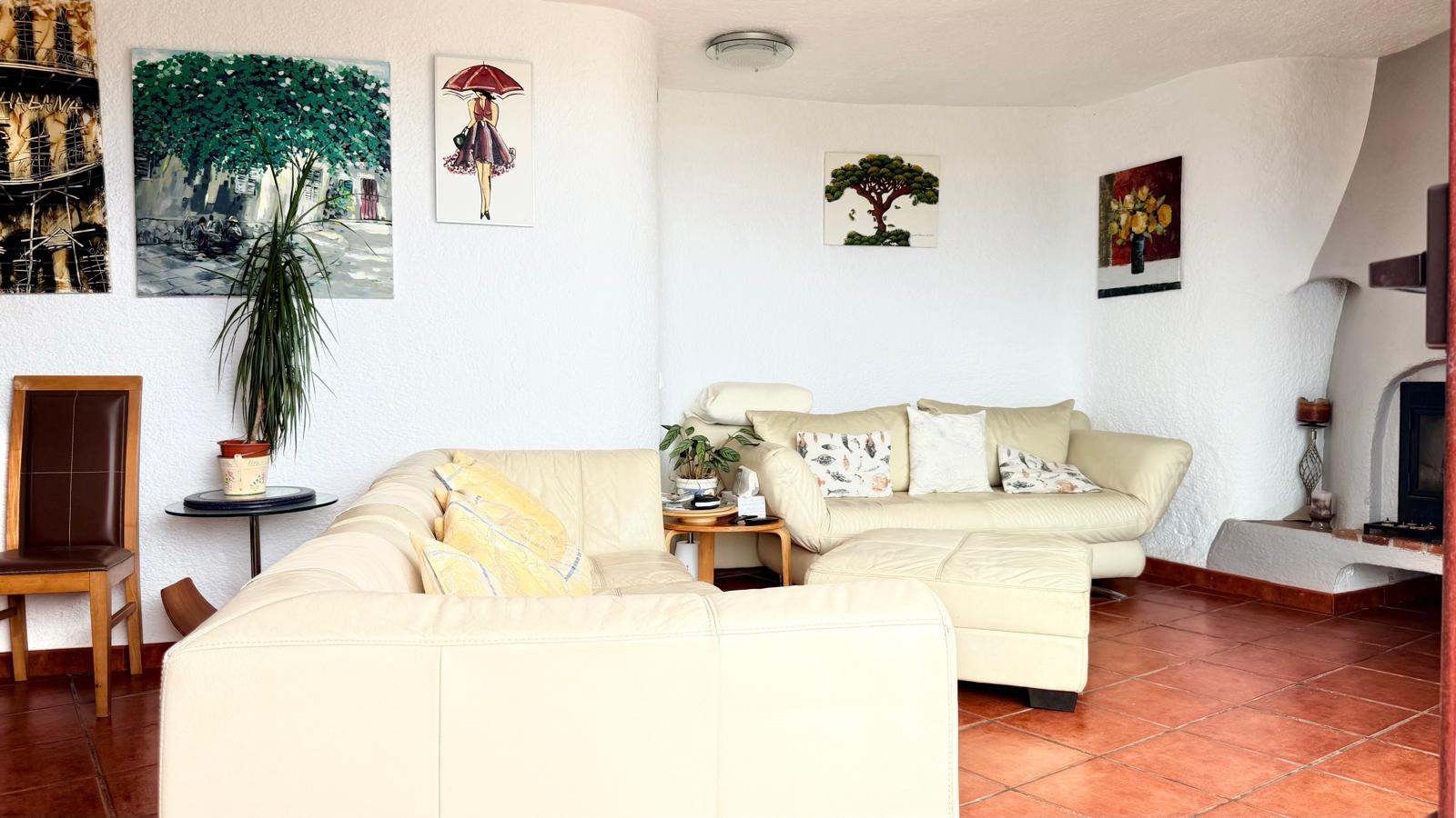 Woonkamer in Ibizastijl met terracotta vloertegels, witte textuurmuren en een houthaard. Ingericht met crème lederen banken, een hocker en wanddecoratie.