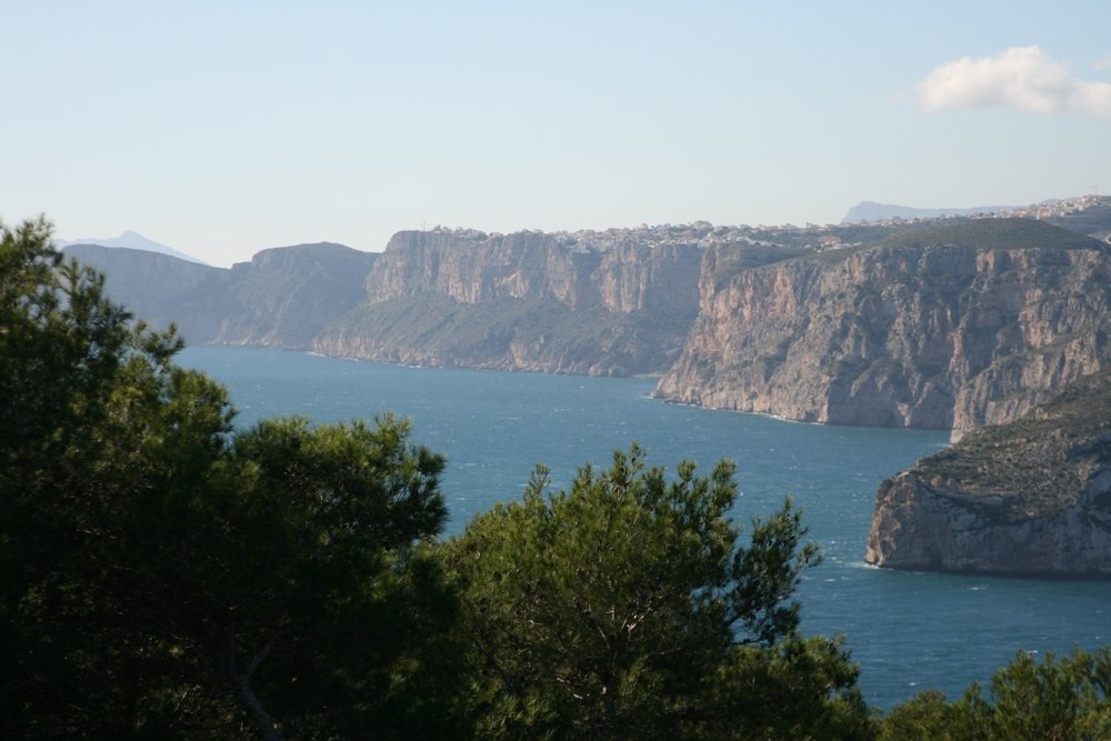 Panoramisch uitzicht op de Middellandse Zee en de kustkliffen van Jávea, omlijst door inheemse dennenbomen onder een heldere hemel.