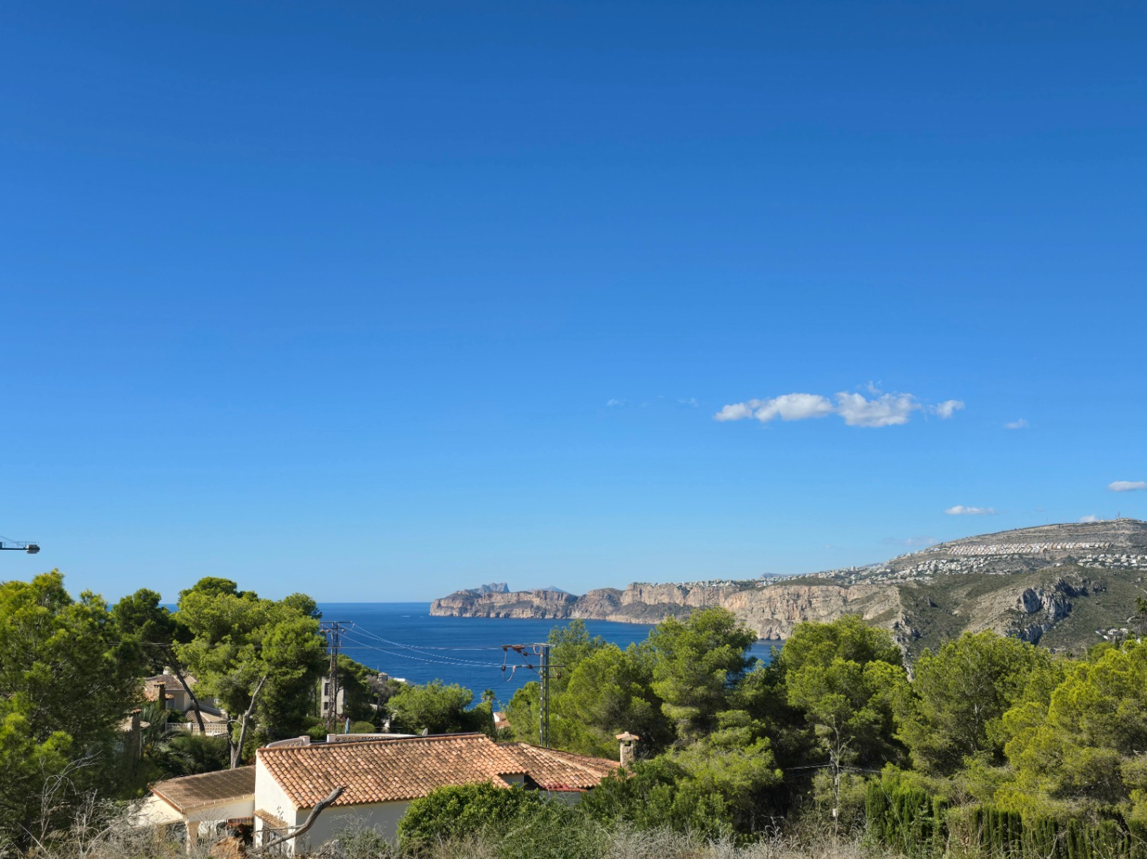 Panoramisch uitzicht op de Middellandse Zee en de kustlijn van Jávea, omgeven door pijnbomen en traditionele daken onder een helderblauwe lucht.