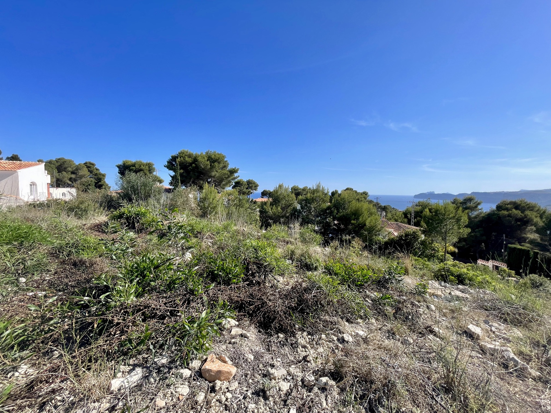 Verhoogd zicht op het perceel met mediterrane vegetatie en pijnbomen, met uitzicht op de zee en de kustlijn van Jávea onder een strakblauwe lucht.