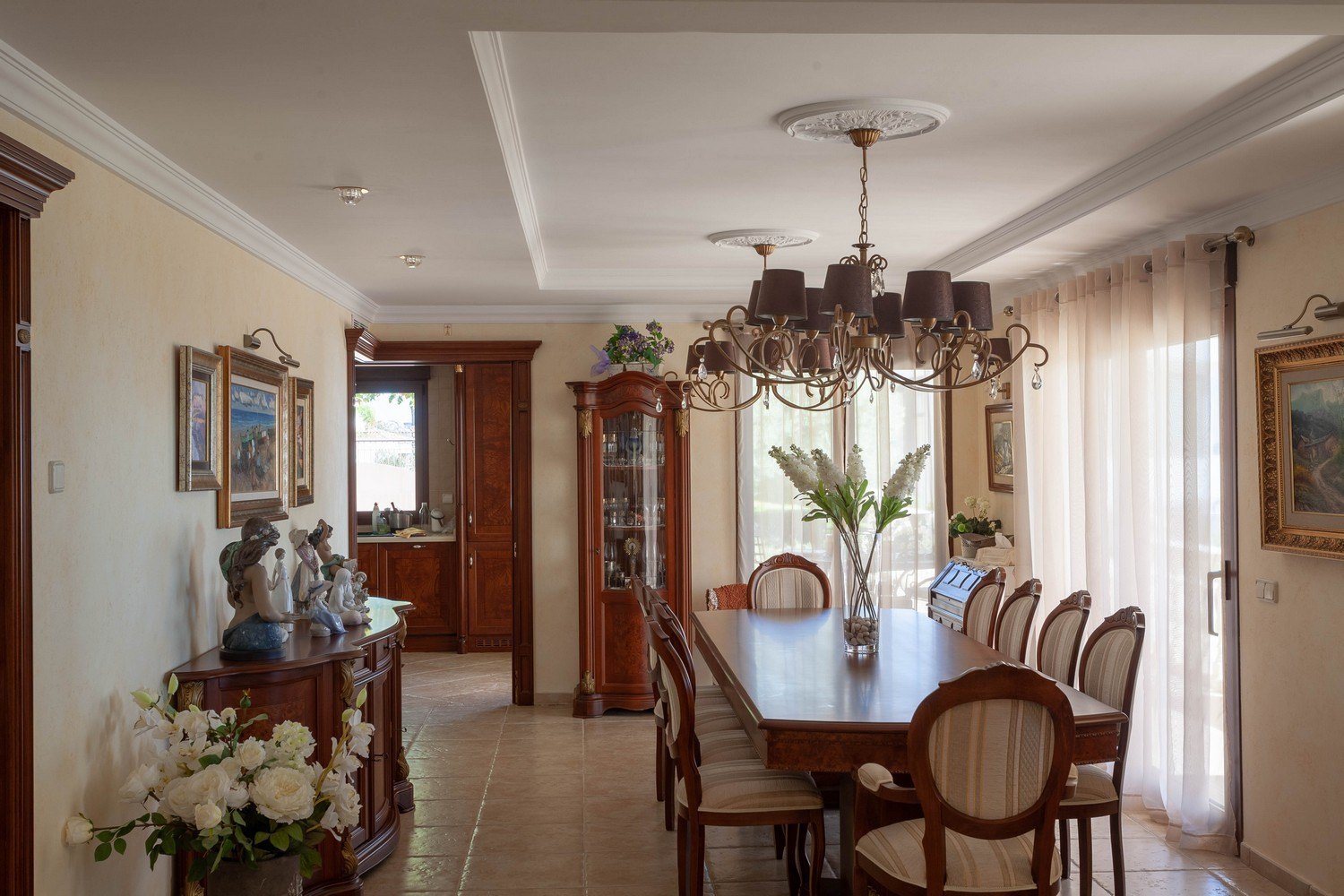 Klassieke eetkamer met houten tafel, kroonluchter en houten vitrinekast met gouden accenten.