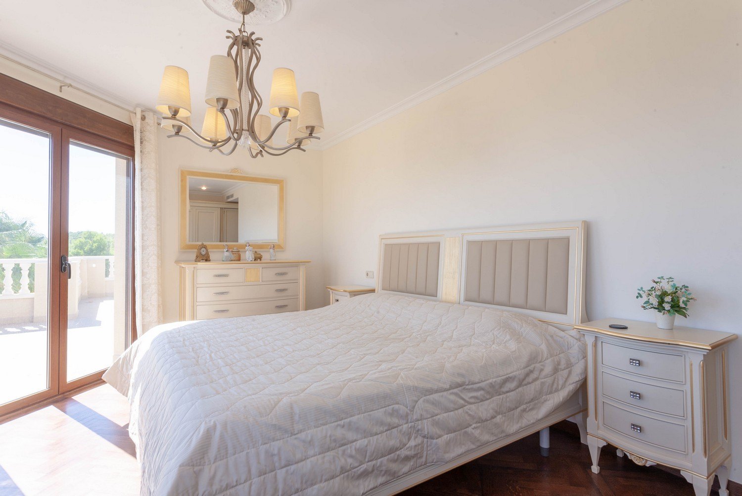 Lichte slaapkamer met tweepersoonsbed, nachtkastjes en een klassieke commode met een goudomrande spiegel.