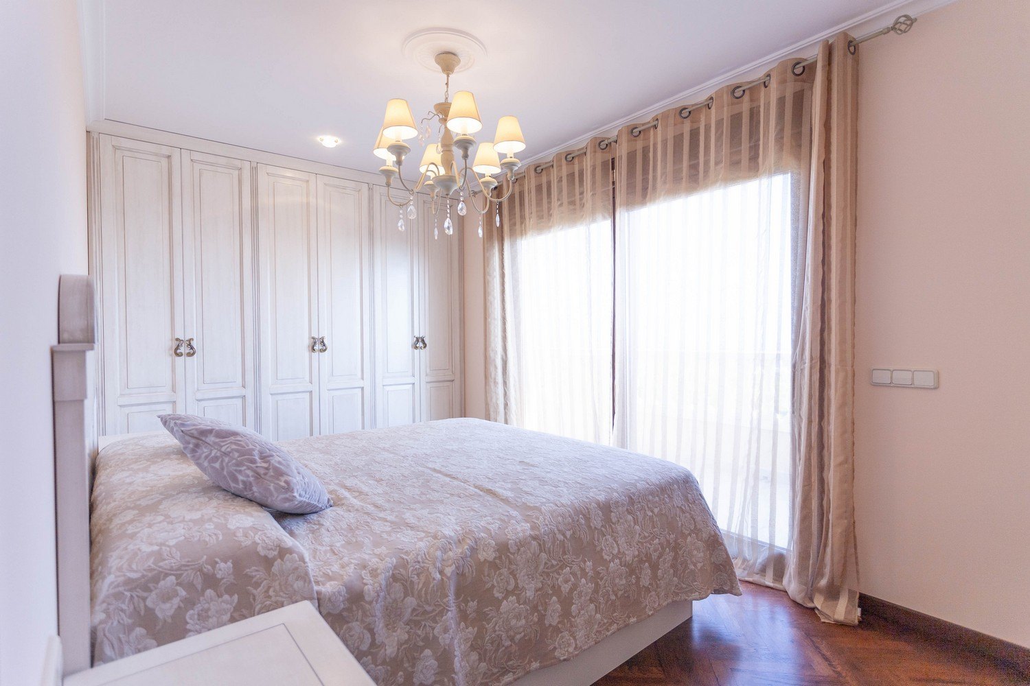 Slaapkamer met tweepersoonsbed, witte inbouwkast, kroonluchter en beige gordijnen.