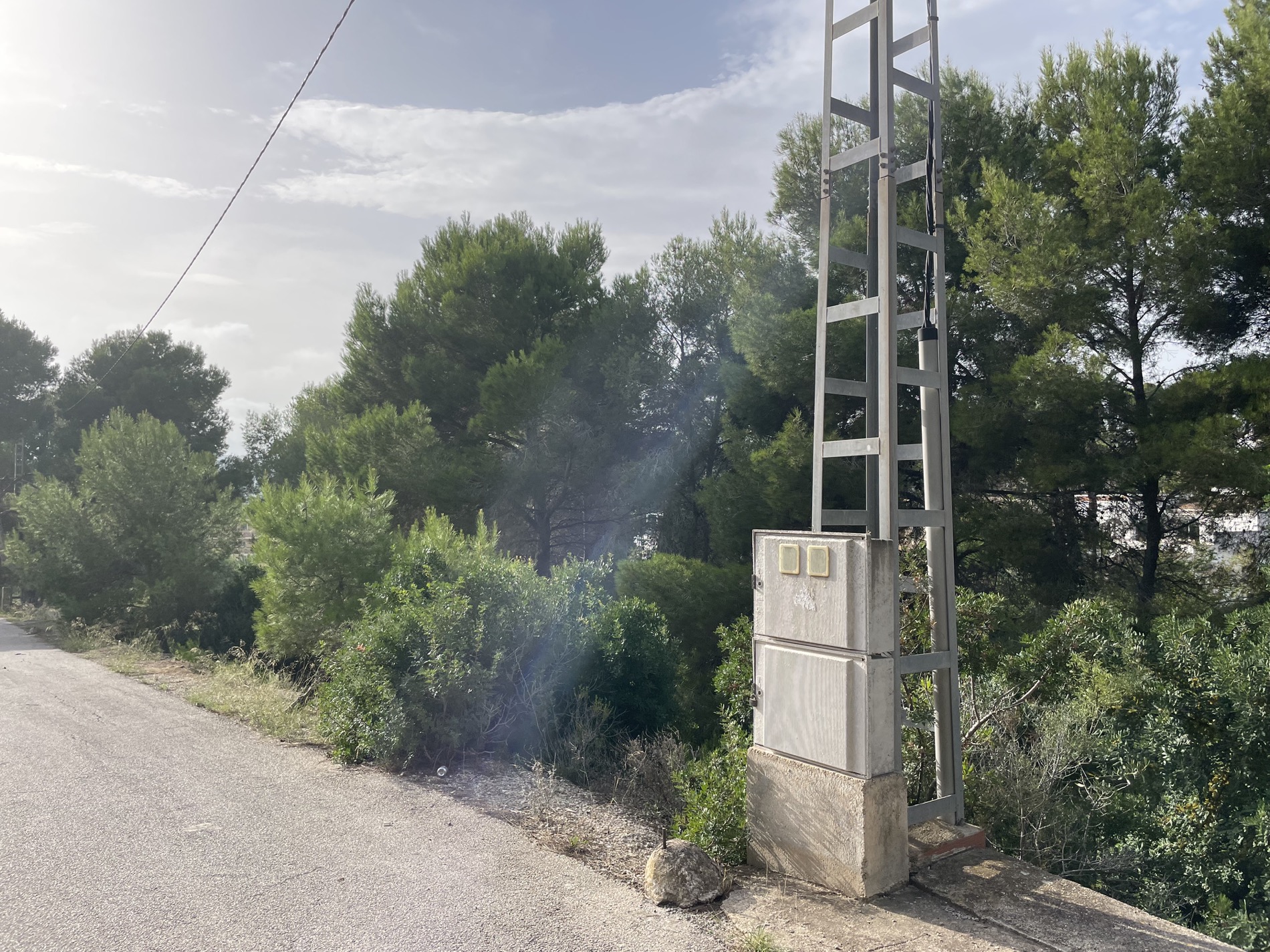 Metalen elektriciteitsmast met aansluitkast langs de geasfalteerde toegangsweg, omgeven door dichte mediterrane begroeiing en pijnbomen.