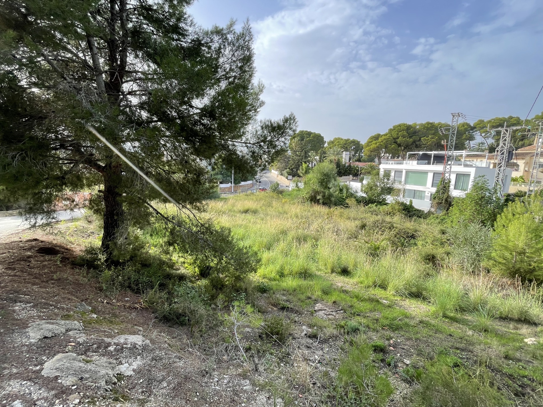 Verhoogd bouwperceel met natuurlijke begroeiing en pijnbomen, grenzend aan een moderne woonwijk in de kuststreek van Moraira.