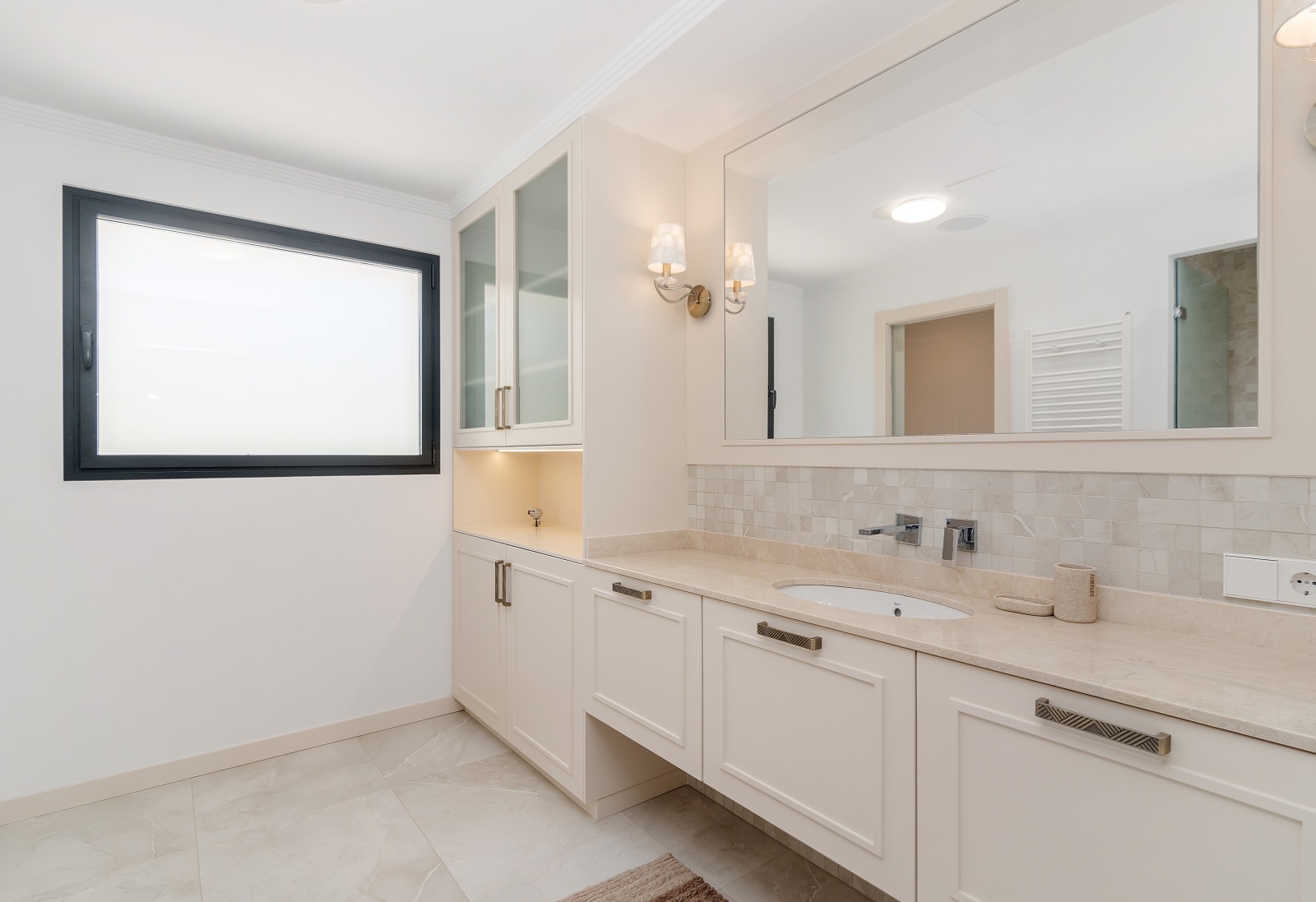 Elegante badkamer met stenen werkblad, geïntegreerde wastafel en crèmekleurige kasten. Voorzien van glazen vitrinekasten met verlichting en klassieke wandlampen.