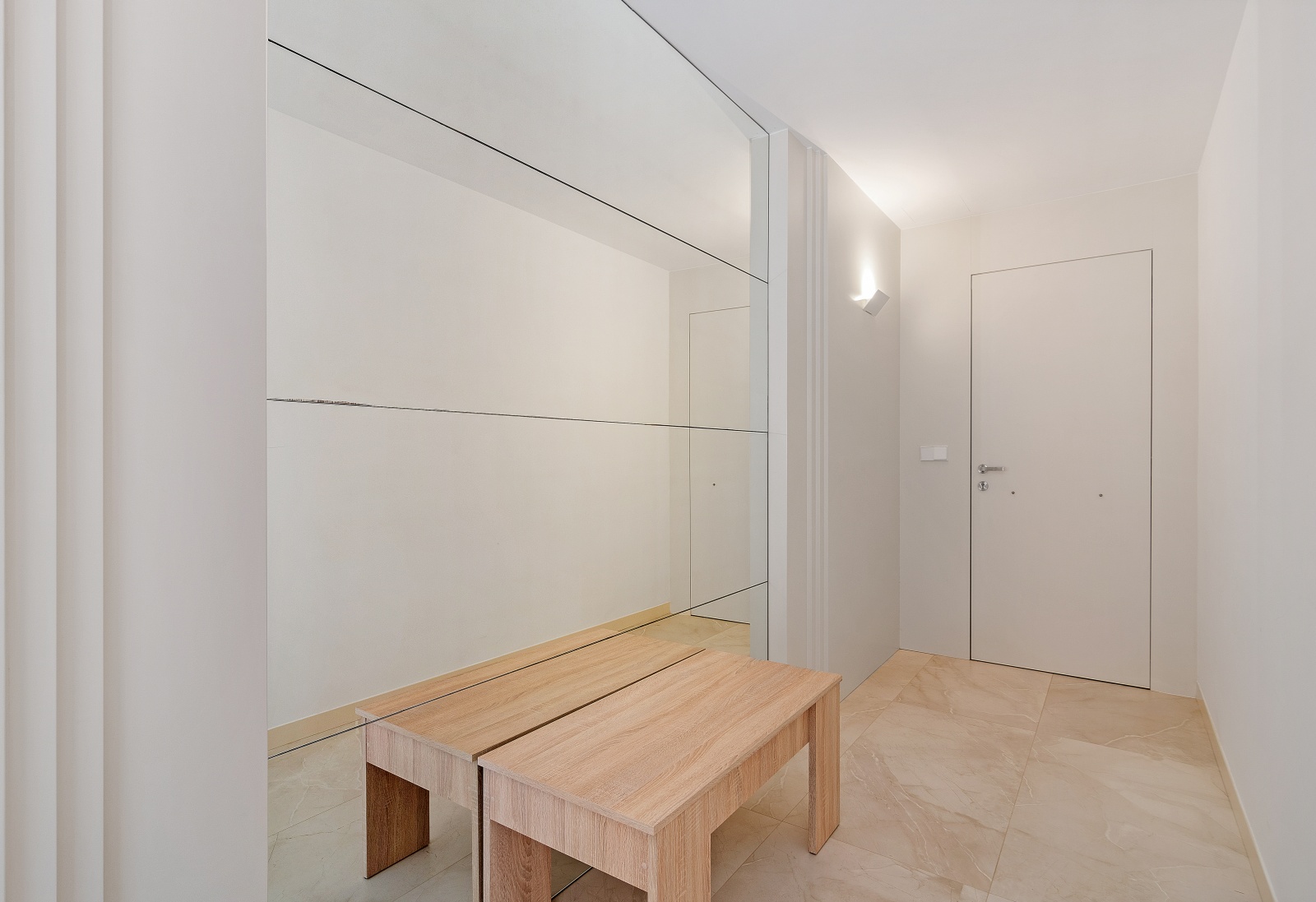 Entreehal met een grote wandspiegel, een licht houten bankje en crème porseleinen vloertegels. Minimalistisch ontwerp met een strakke witte deur en wandverlichting.