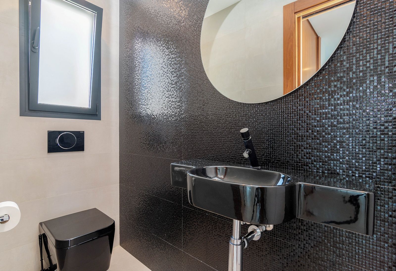 Gastentoilet met een zwarte mozaïekwand, een bijpassende zwarte wastafel en een hangend toilet. Voorzien van een ronde spiegel en een raam met melkglas.