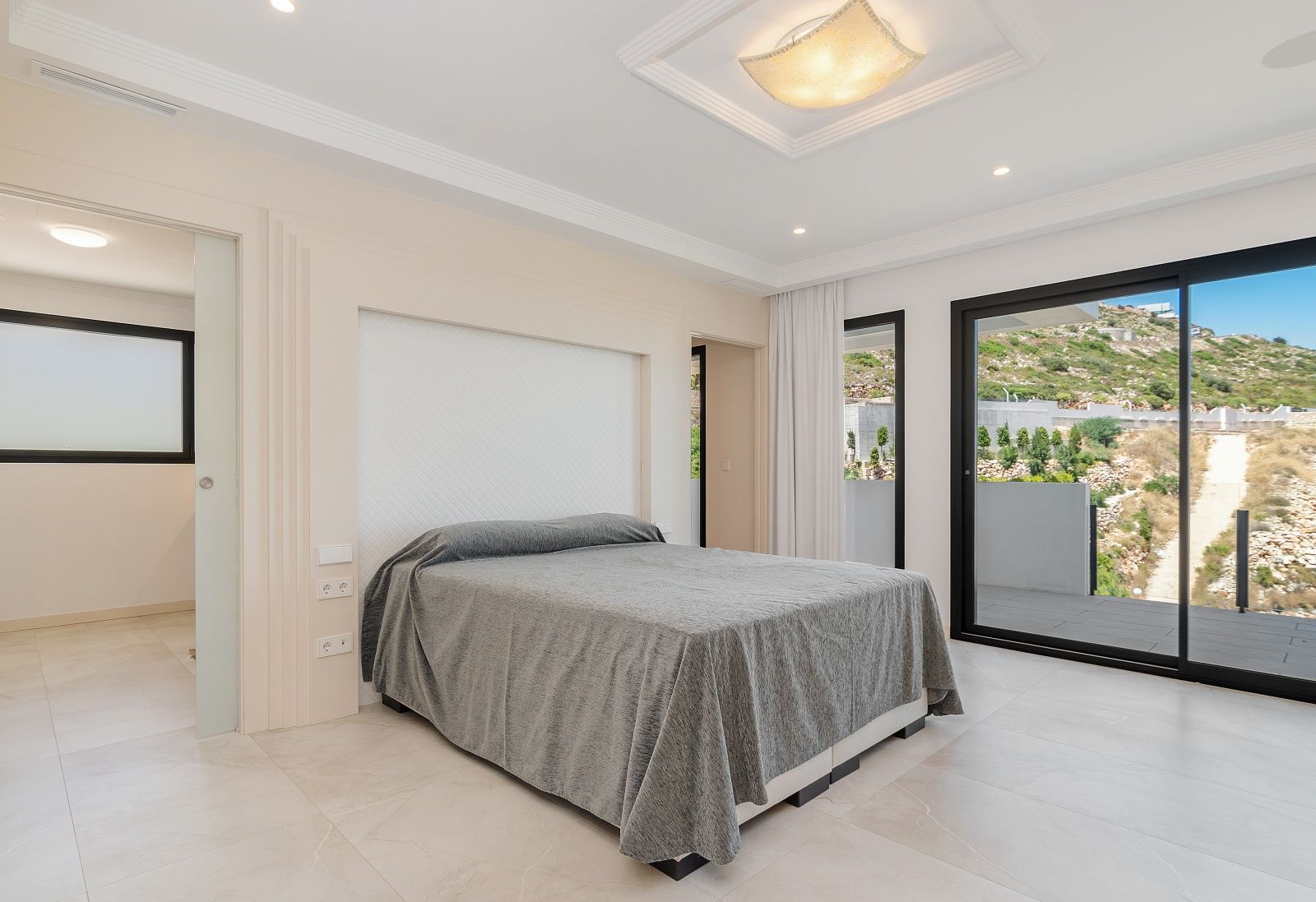 Lichte slaapkamer met grote glazen schuifdeuren naar het terras, een geïntegreerde hoofdbordnis en elegante inbouwverlichting in het plafond.