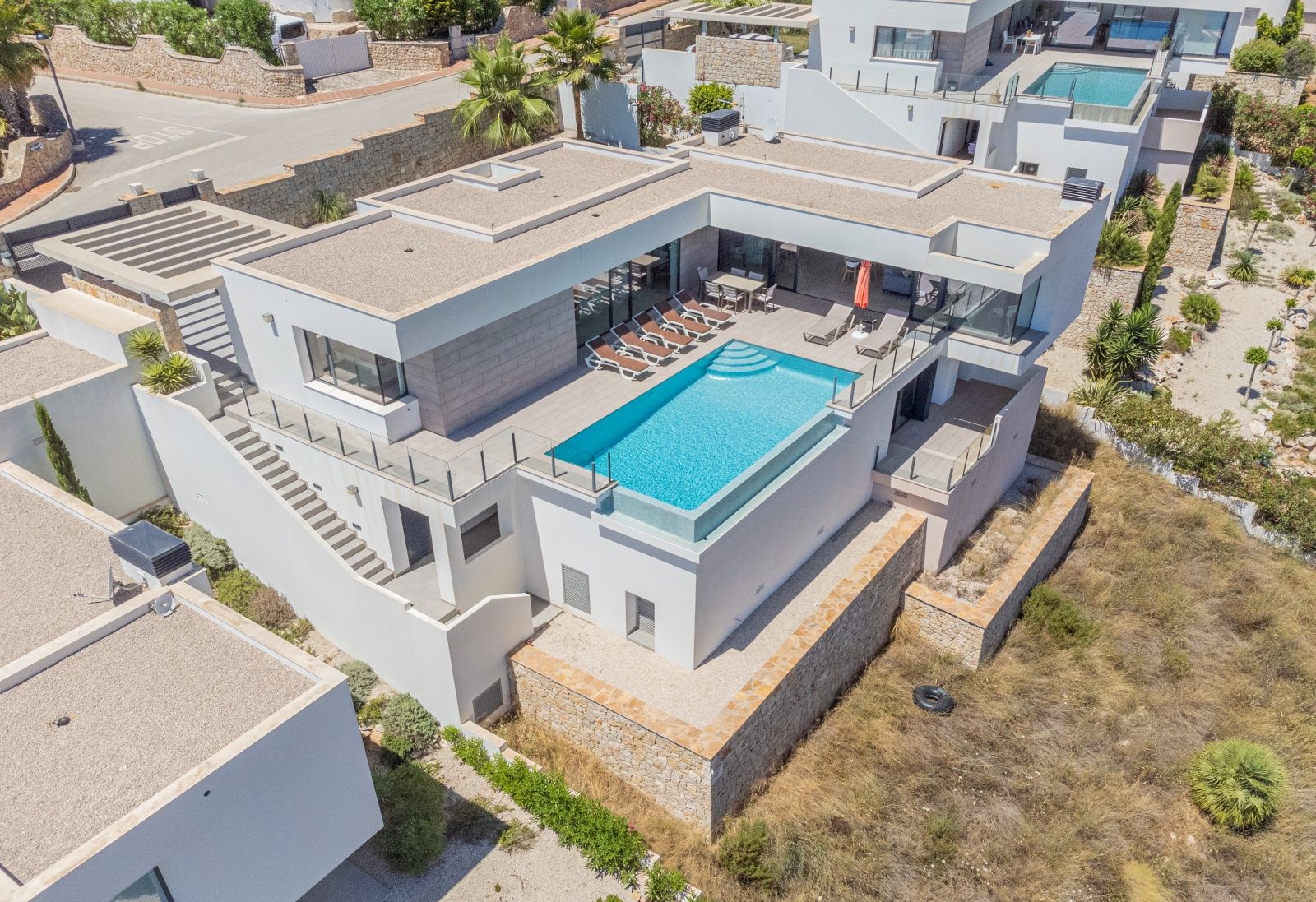 Luchtfoto van de moderne villa met overloopzwembad met glazen wand, ruim zonneterras en minimalistische witte architectuur geïntegreerd in het terrein.