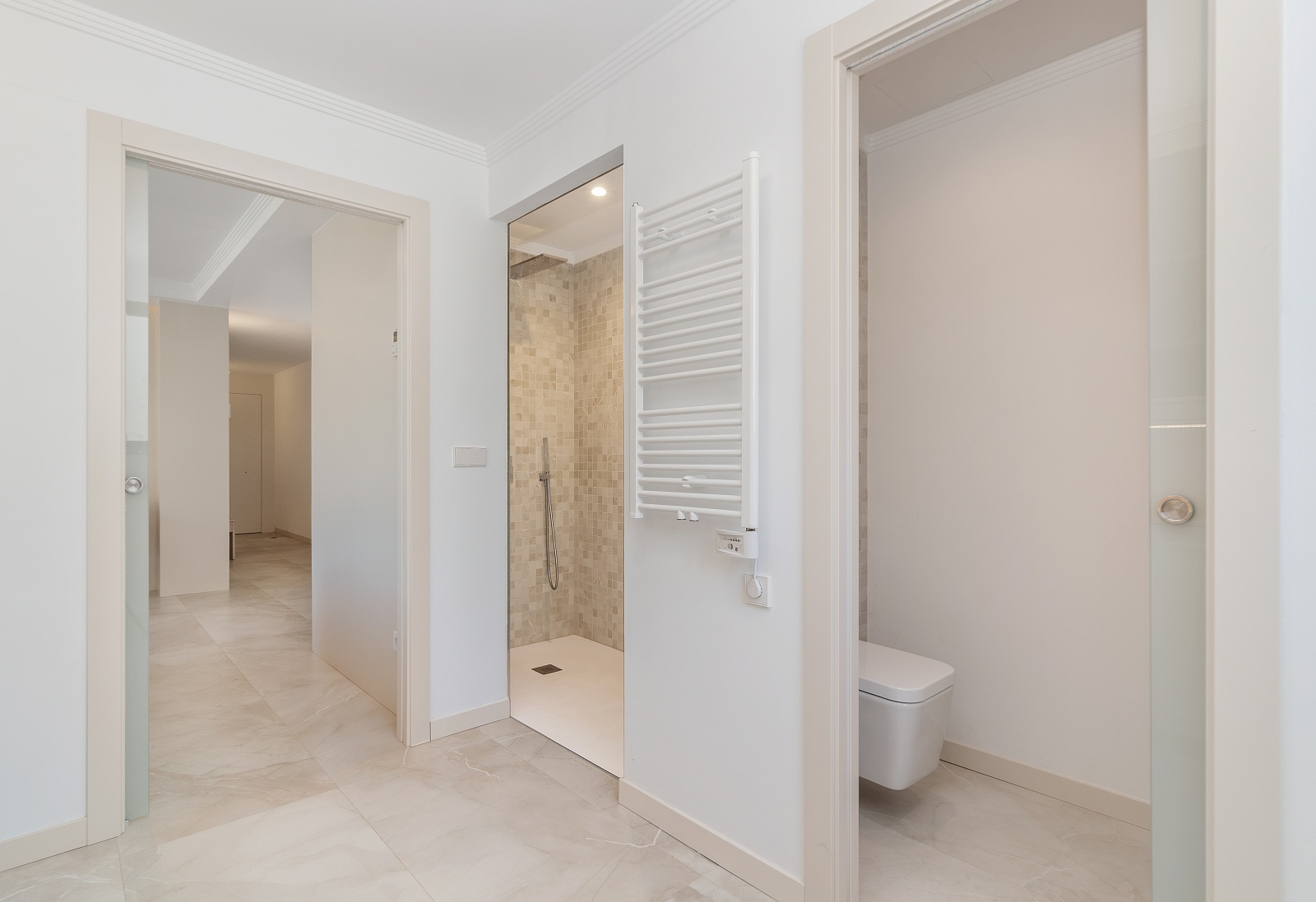 Moderne badkamer met inloopdouche in mozaïek, hangend toilet, witte handdoekradiator en glazen schuifdeuren op marmerlook vloertegels.