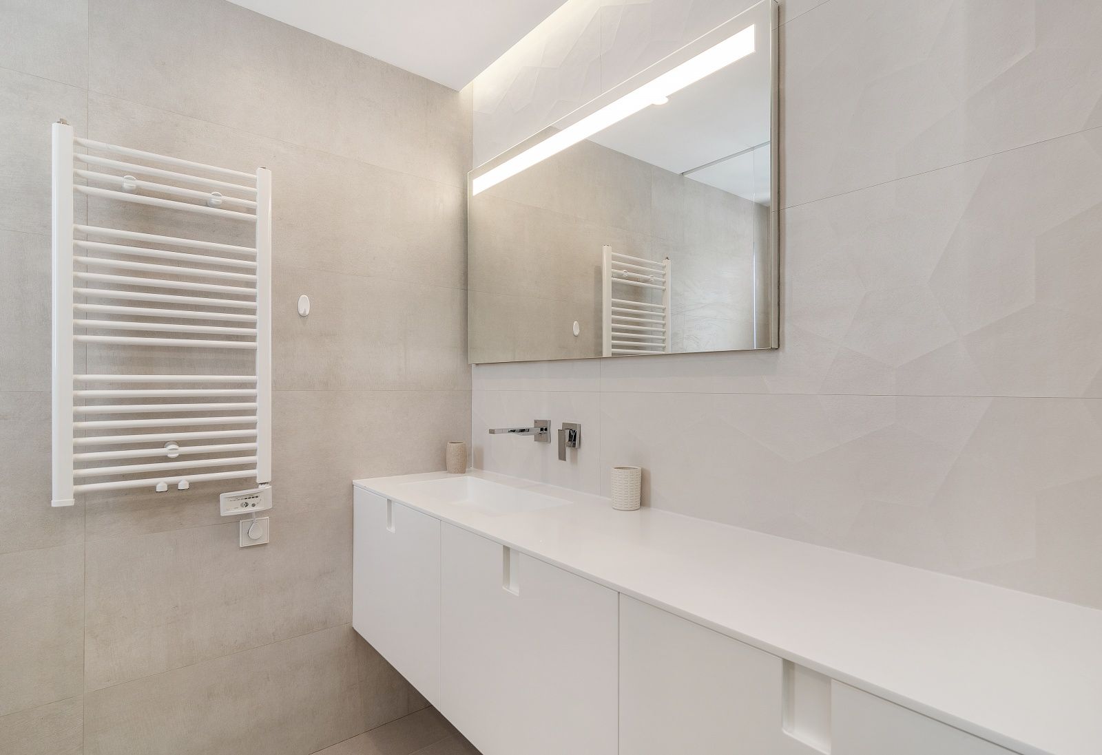 Moderne badkamer met wit zwevend wastafelmeubel, inbouwkranen, grote spiegel met LED-verlichting en een witte handdoekradiator tegen getextureerde wandtegels.