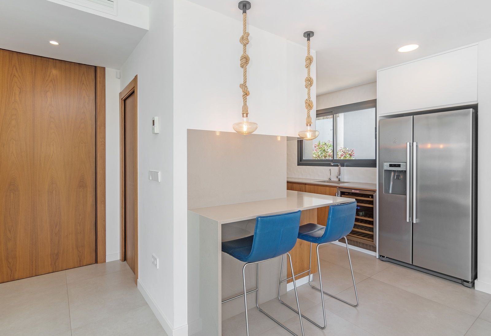 Moderne keuken met een grijs ontbijtbar, blauwe krukken en decoratieve touwhanglampen. Voorzien van een Amerikaanse koelkast en ingebouwde wijnkoeler.