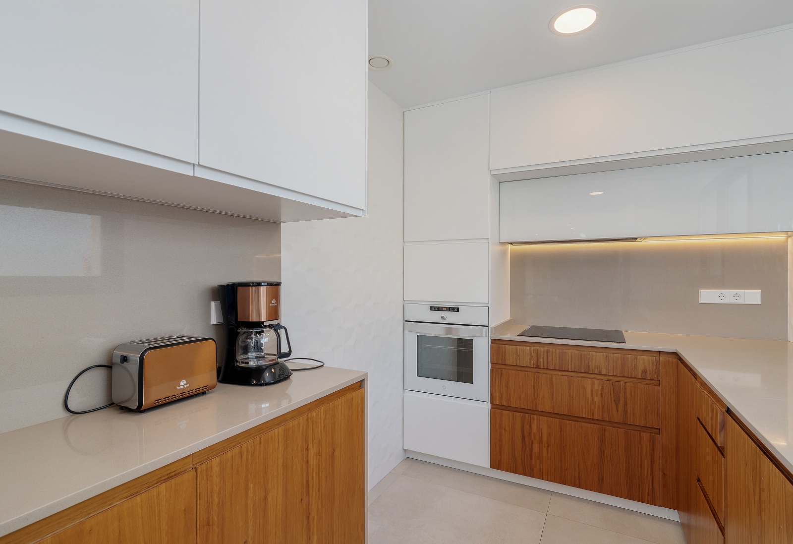 Moderne keuken met witte bovenkasten, houten onderkasten, ingebouwde oven, inductiekookplaat en een kwarts werkblad met subtiele LED-verlichting.