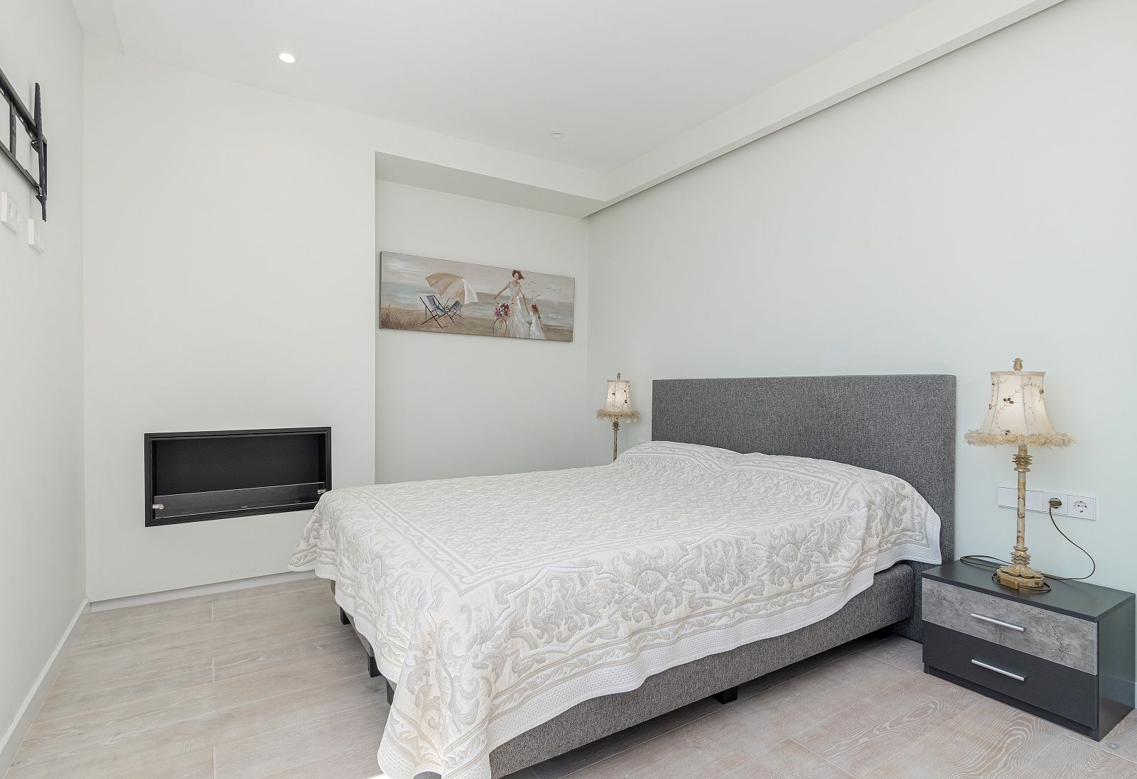 Moderne slaapkamer met ingebouwde haard, grijs stoffen hoofdbord, design nachtkastje en lichte keramische vloer met houtlook.