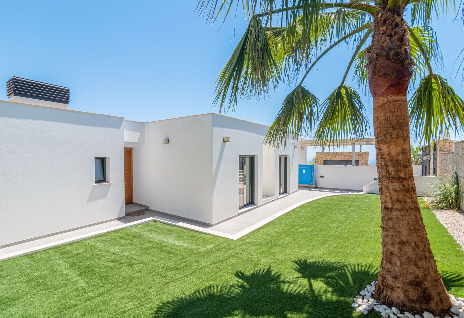 Moderne witte villa met een strak kunstgrasveld, een palmboom op de voorgrond en een houten voordeur in een zonnige setting.