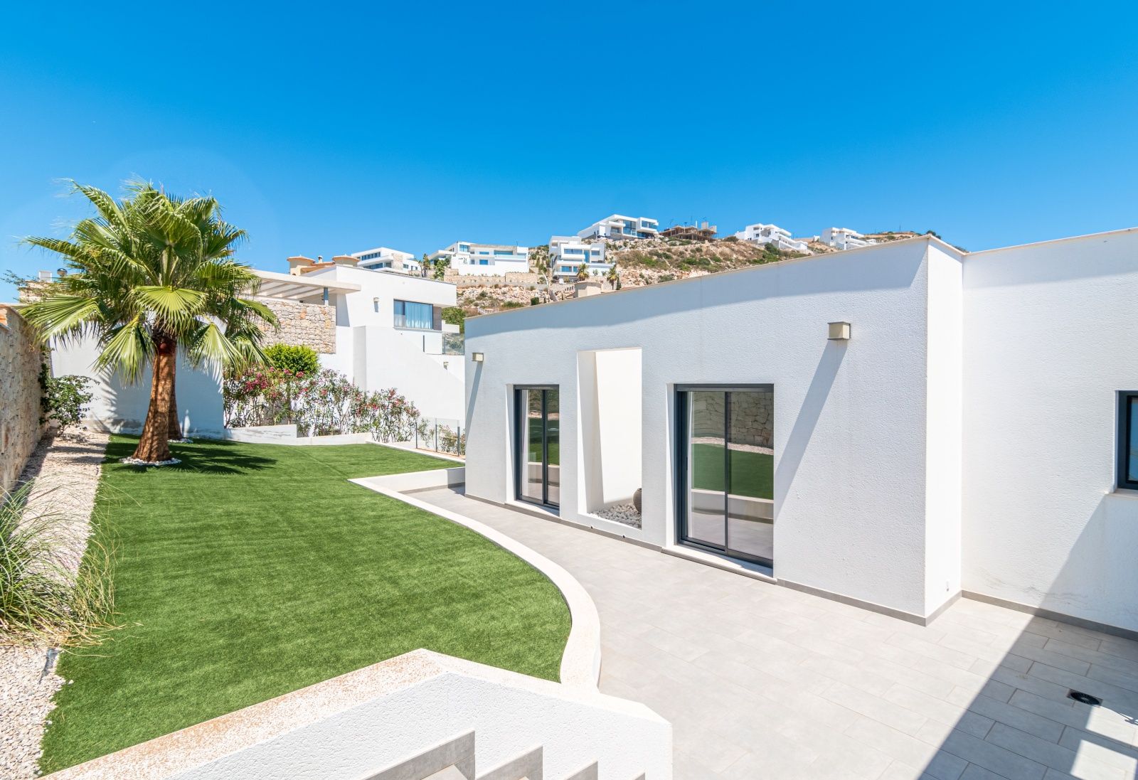 Moderne witte villa met grote glazen schuifdeuren, een kunstgrasveld met palmboom en een betegeld terras in een residentiële omgeving.