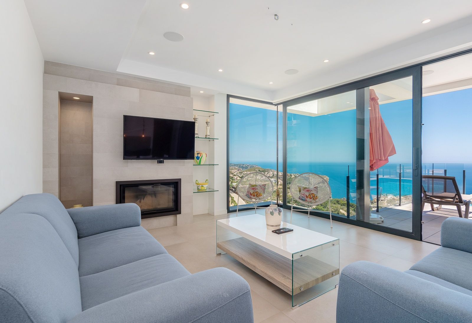 Moderne woonkamer met blauwe banken, een ingebouwde haard en grote glazen schuifdeuren met panoramisch uitzicht op de Middellandse Zee.