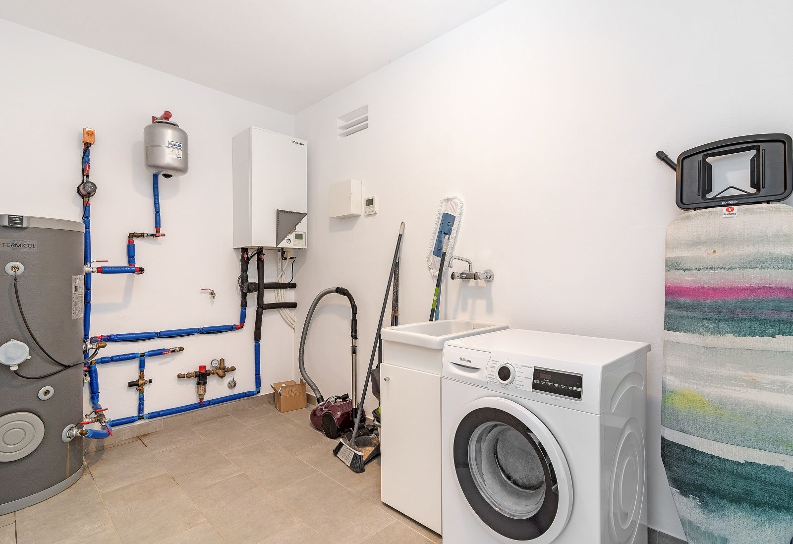 Praktische wasruimte en technische ruimte voorzien van een wasmachine, uitstortgootsteen en zichtbare leidingen voor verwarming en sanitair.