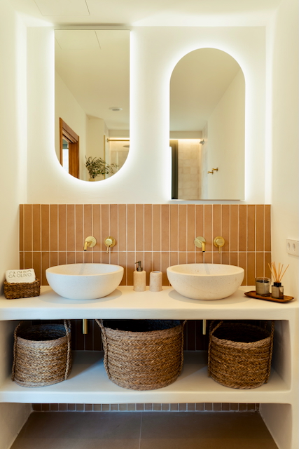 Badkamer met dubbele stenen waskommen, gouden inbouwkranen en verticale terracotta tegels. Voorzien van verlichte boogspiegels en rieten opbergmanden.