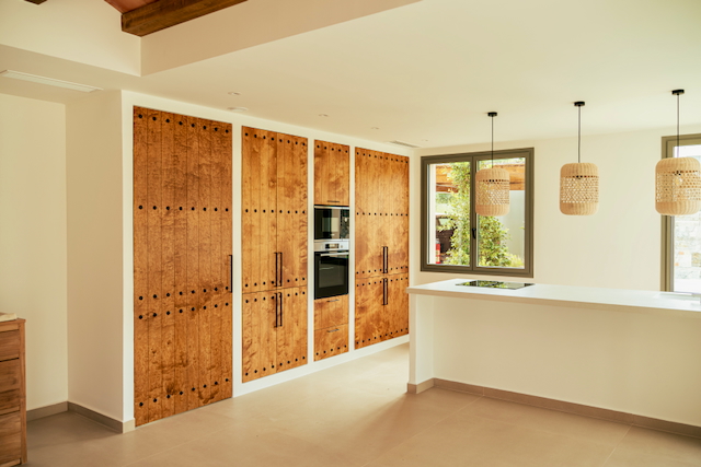 Designkeuken met rustieke houten kasten en geïntegreerde apparatuur. Een wit kookeiland met inductiekookplaat wordt verlicht door drie geweven hanglampen.