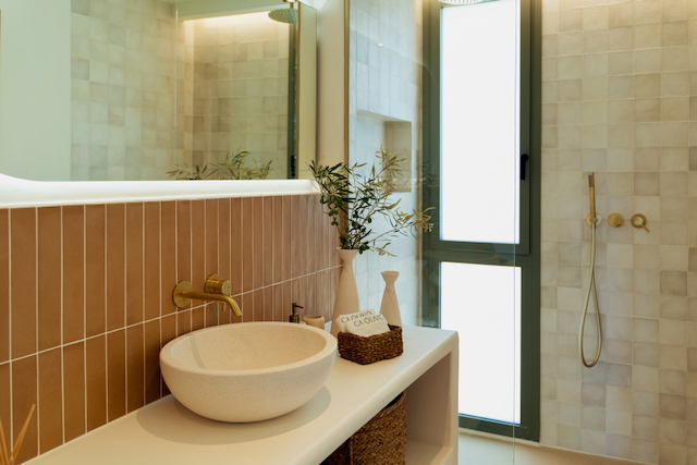 Moderne badkamer met een stenen opzetwastafel, geborsteld gouden kranen en verticale tegels. Voorzien van een inloopdouche met glazen wand en natuurlijk licht.
