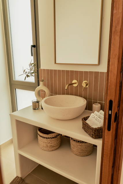 Moderne badkamer met een stenen opzetwastafel, goudkleurige inbouwkranen en verticale terracotta tegels boven een wit gestuct meubel.