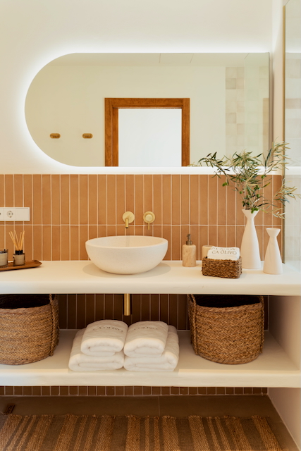 Moderne badkamer met verticale terracotta tegels, een opzetwastafel en goudkleurige kranen. Voorzien van een verlichte spiegel en open planken met rieten manden.