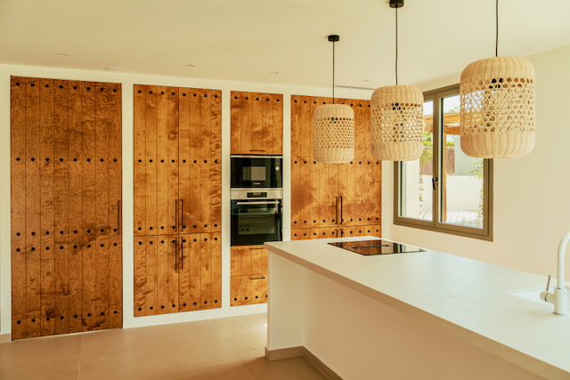 Open keuken met wit kookeiland, houten kastenwanden voorzien van siernagels en sfeervolle gevlochten hanglampen boven het werkblad.