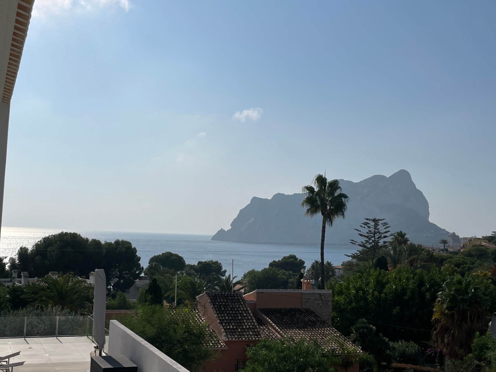 Panoramisch uitzicht op de Peñón de Ifach en de Middellandse Zee vanaf een verhoogd terras met glazen balustrade en weelderige kustvegetatie.