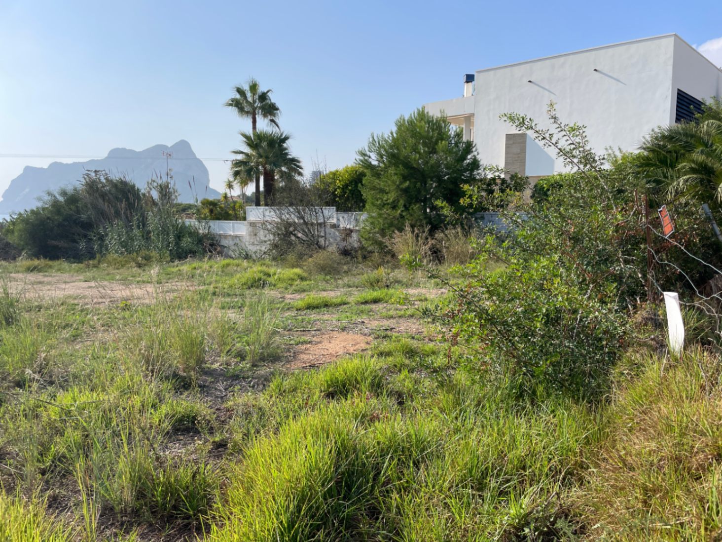 Vlak bouwperceel in Bassetes, Calpe, met uitzicht op de Peñón de Ifach. Het terrein is omgeven door natuurlijke begroeiing en een moderne witte villa.
