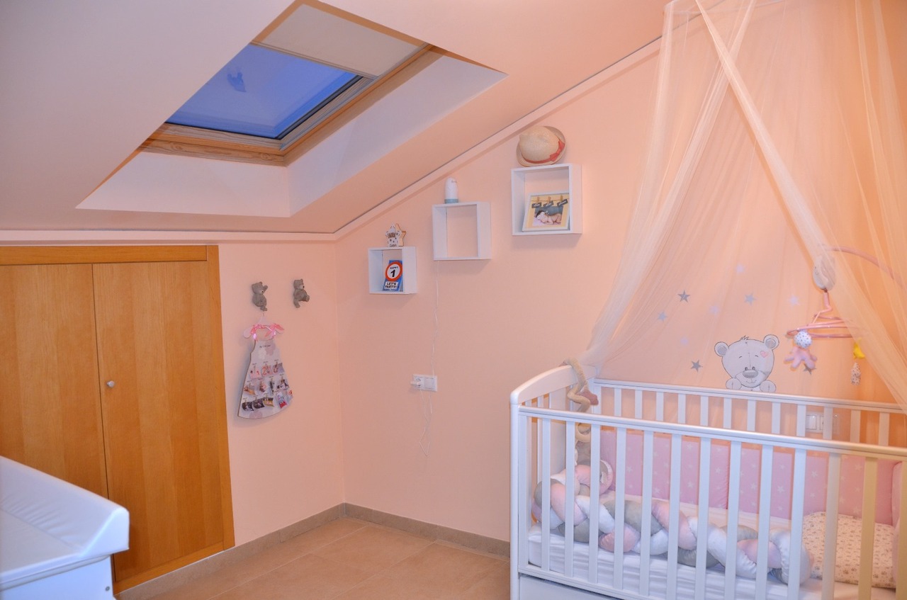 Babykamer met schuin dak en dakraam, wit ledikant met hemeltje, roze wanden en een ingebouwde houten kledingkast.