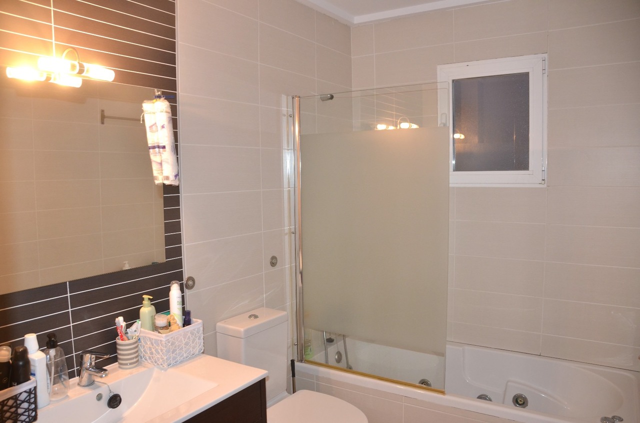 Moderne badkamer met wit wastafelmeubel, een ligbad met matglazen douchescherm en beige wandtegels gecombineerd met een donkere accentwand achter de spiegel.