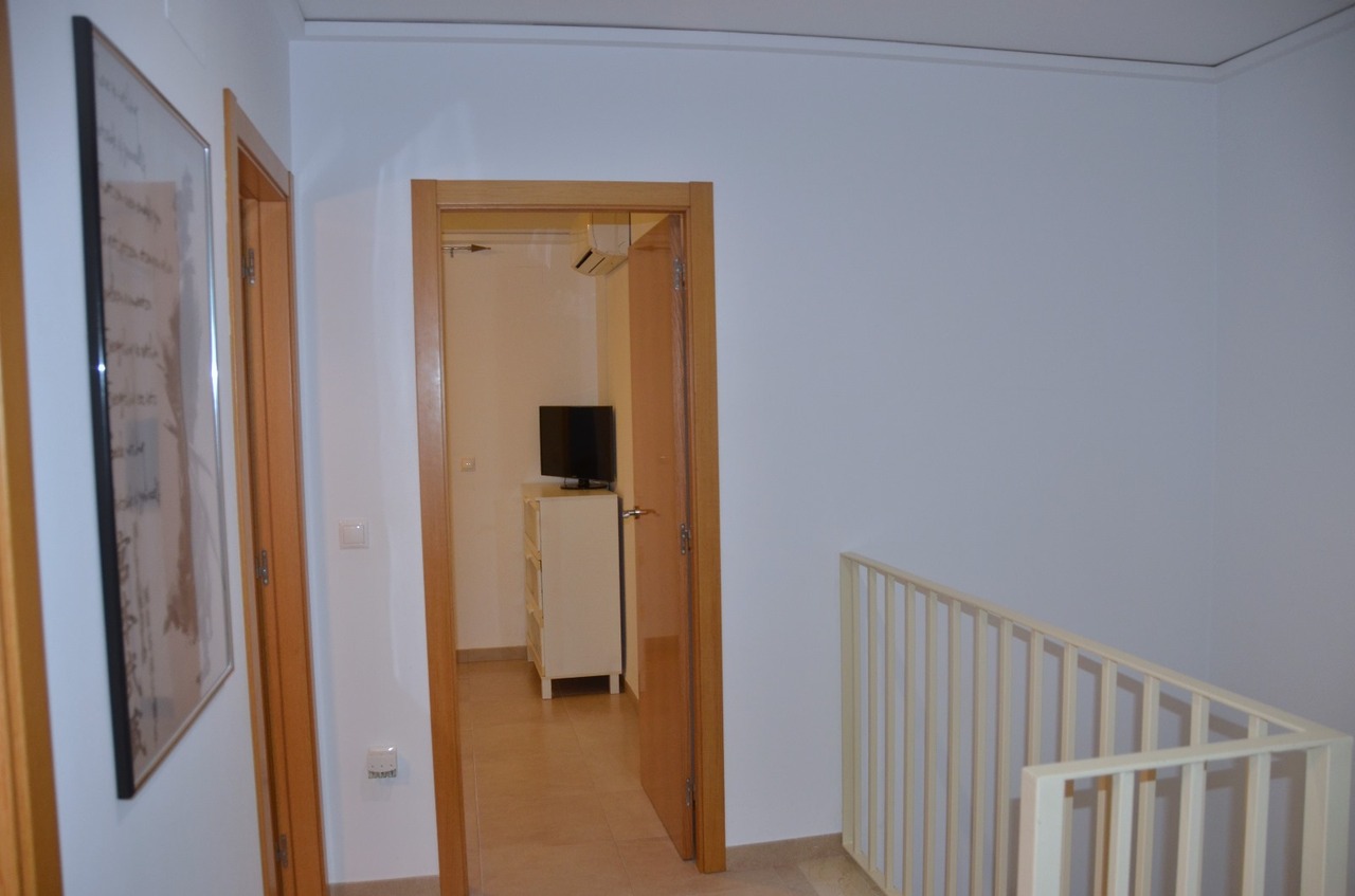 Overloop op de bovenverdieping met licht houten deurkozijnen, een witte metalen trapleuning en doorkijk naar een slaapkamer met witte ladekast en tv.