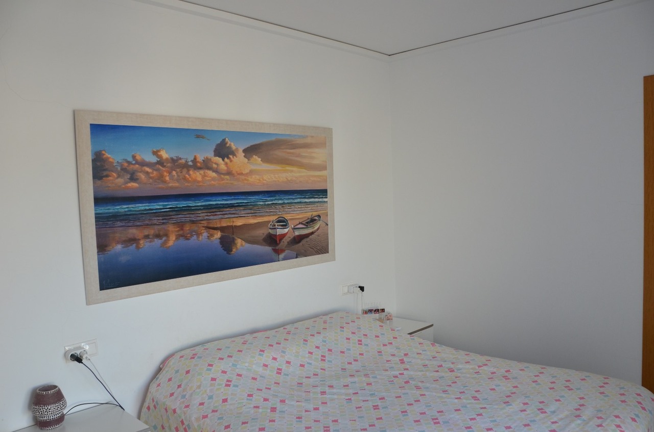 Slaapkamer met een groot schilderij van een kustlandschap boven het bed, witte muren en een sprei met een kleurrijk geometrisch patroon.