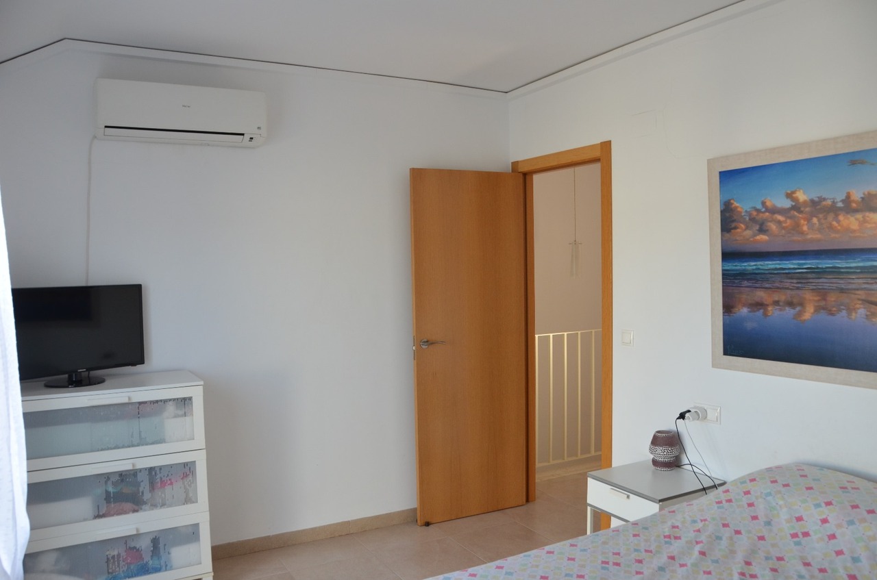 Slaapkamer met split-airconditioning, een witte ladekast met televisie en een houten deur naar de overloop. Voorzien van een tegelvloer en wanddecoratie.