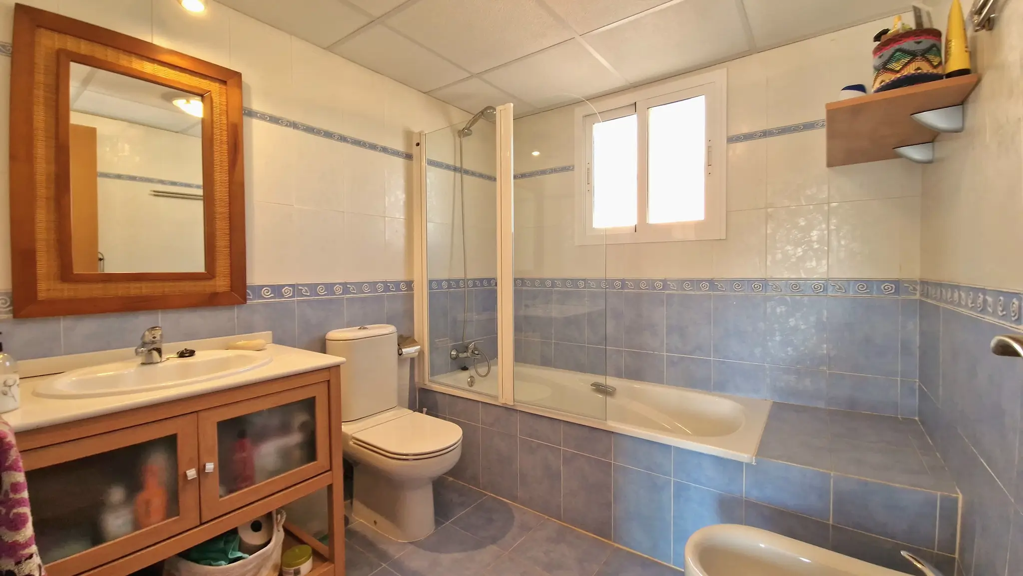 Badkamer met blauw-witte betegeling, een houten wastafelmeubel en een ligbad met glazen douchescherm, voorzien van natuurlijk lichtinval via een raam.