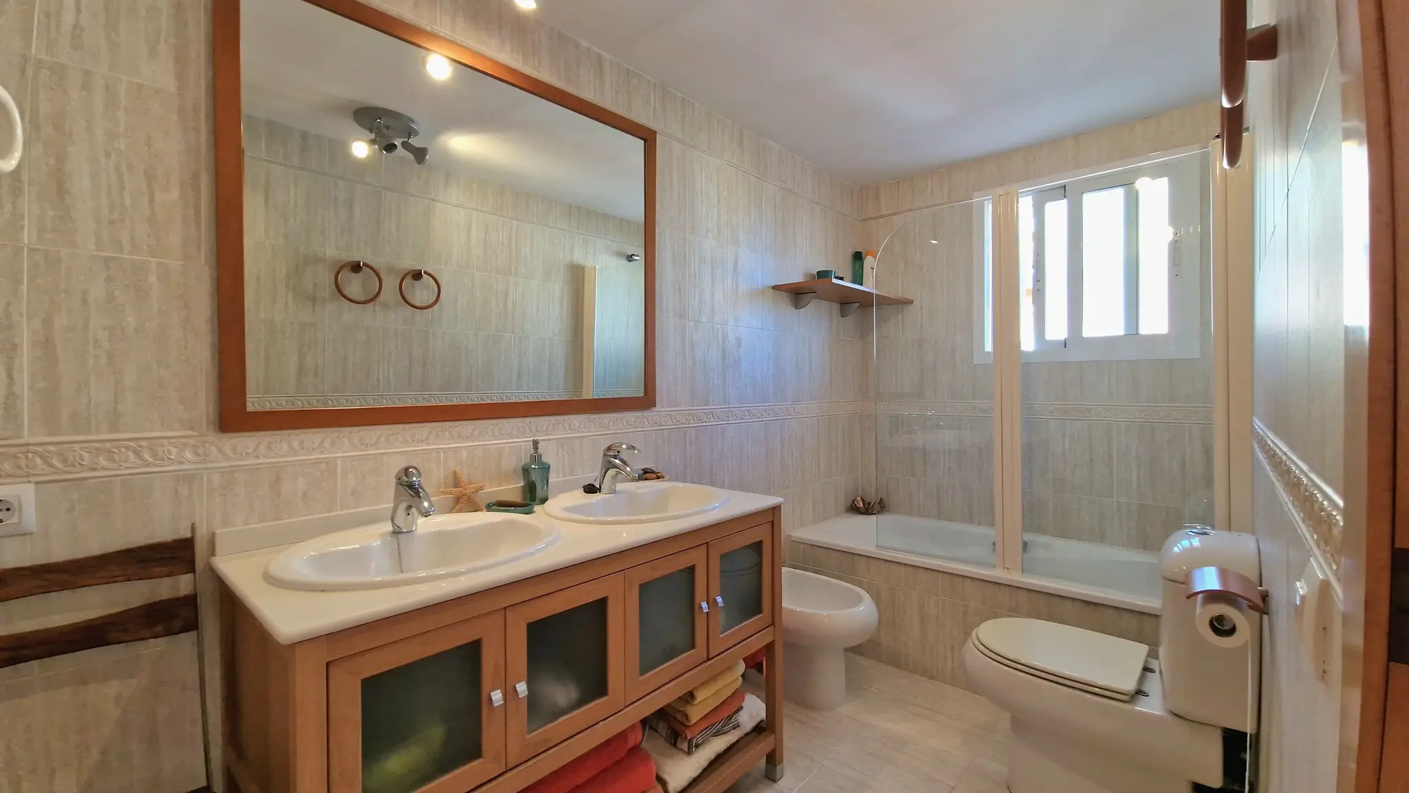 Badkamer met dubbele wastafel op een houten meubel, grote spiegel, bidet en toilet. Voorzien van een ligbad met glazen wand en neutraal tegelwerk.