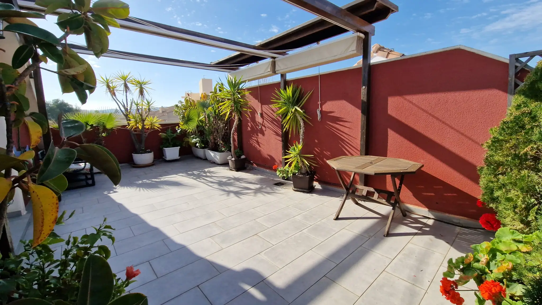 Buitenterras met grijze vloertegels, houten pergola met uitschuifbaar zonnescherm en diverse potplanten tegen een terracotta muur.
