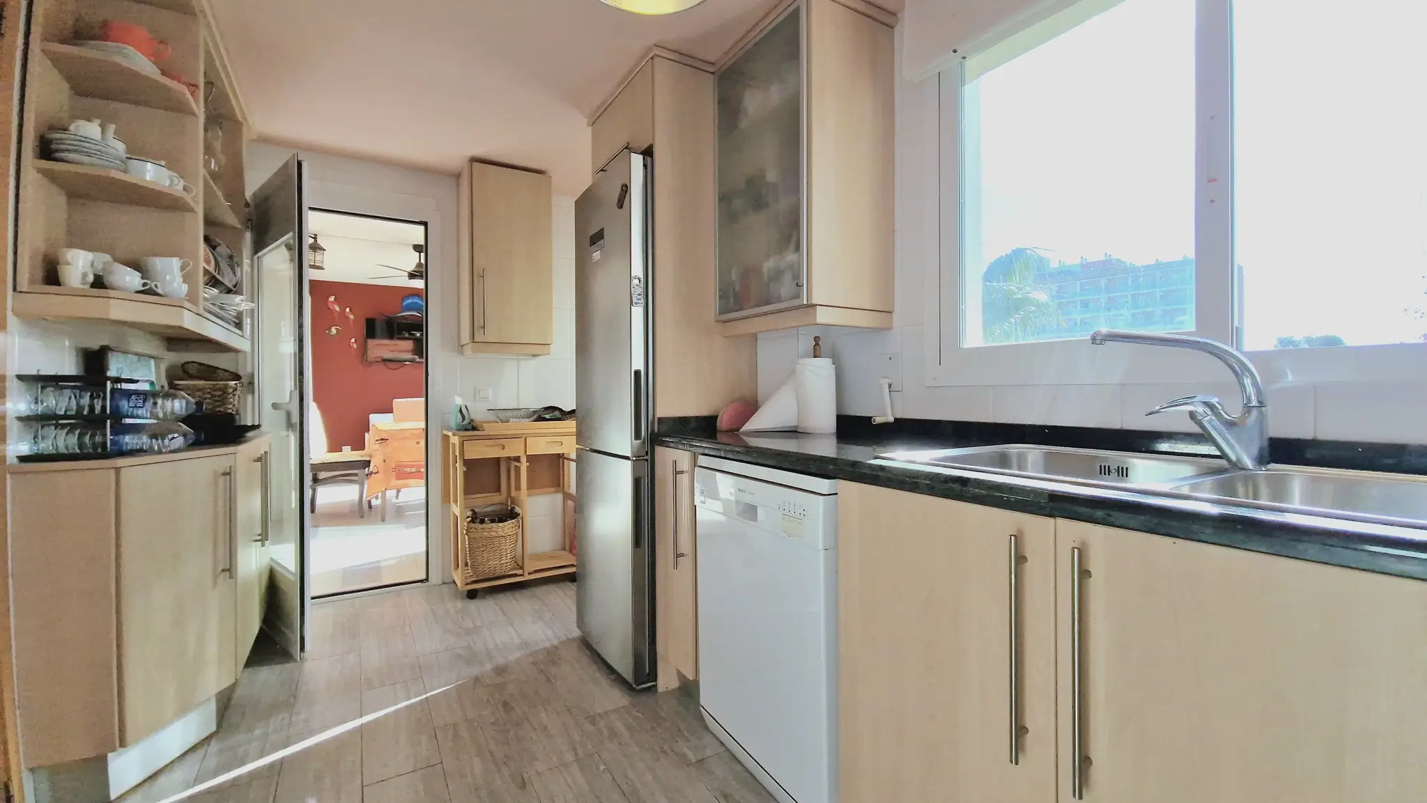 Functionele keuken met lichte houten kasten, donker granieten werkblad, roestvrijstalen spoelbak en een groot raam voor natuurlijke lichtinval.