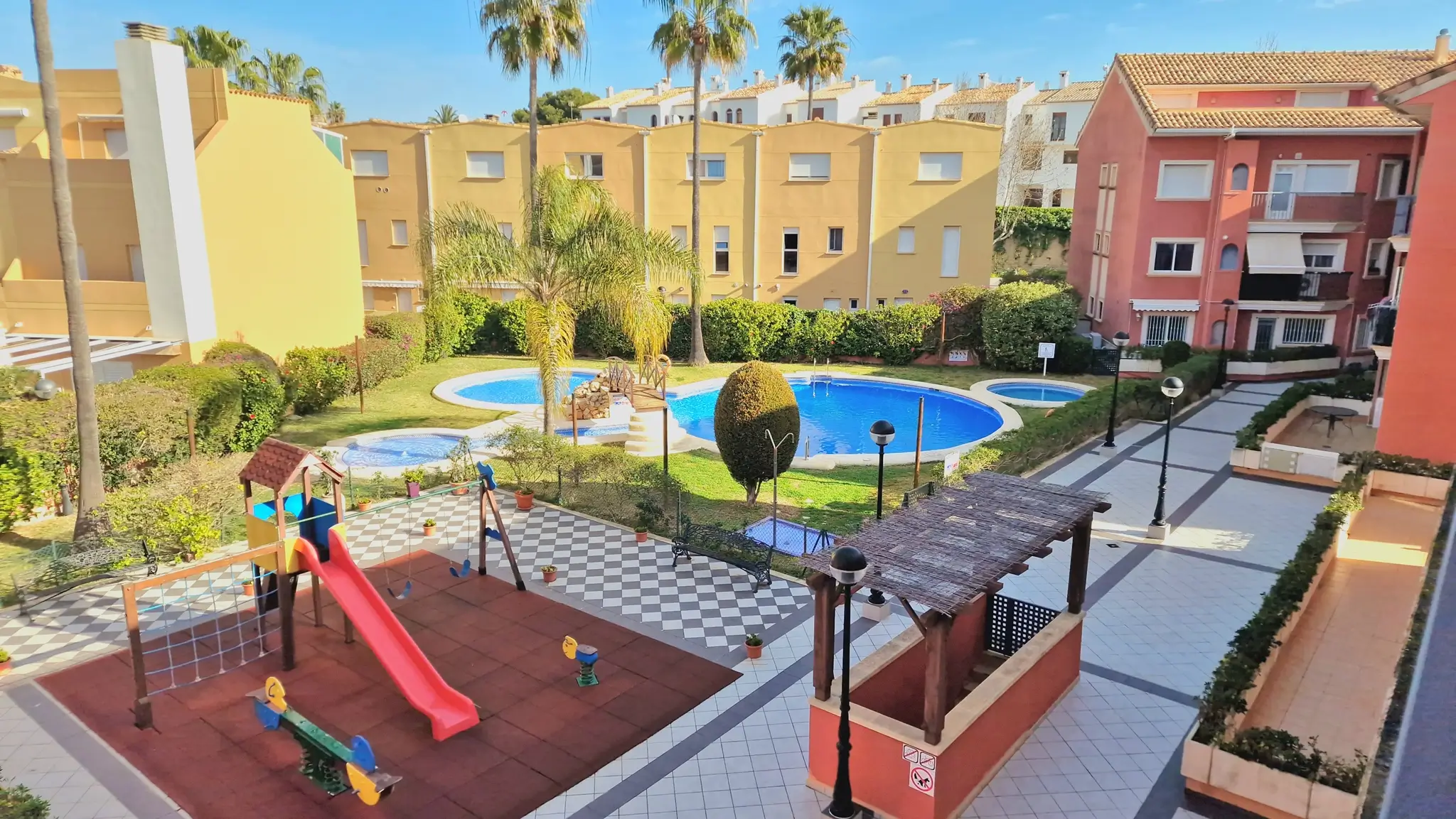 Gemeenschappelijke zone met een kinderspeeltuin, een groot zwembad met bruggetje en verzorgde tuinen, omringd door mediterrane woongebouwen en palmbomen.