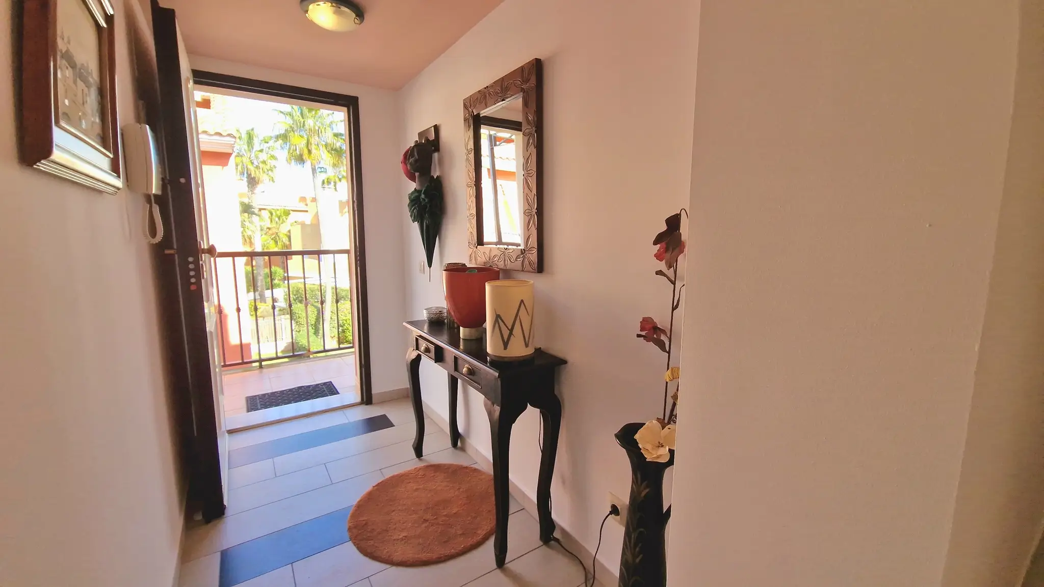 Hal met een donkerhouten consoletafel, decoratieve spiegel en tegelvloer, die uitkomt op een balkon met uitzicht op palmbomen en natuurlijk licht.