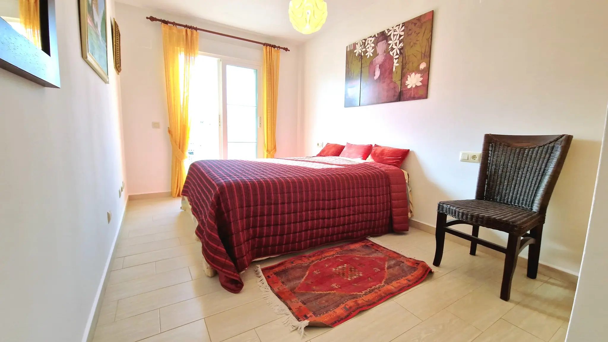 Lichte slaapkamer met tweepersoonsbed, rode gewatteerde sprei, gele gordijnen, een donkere rieten stoel en een decoratief Boeddha-triptiek aan de muur.